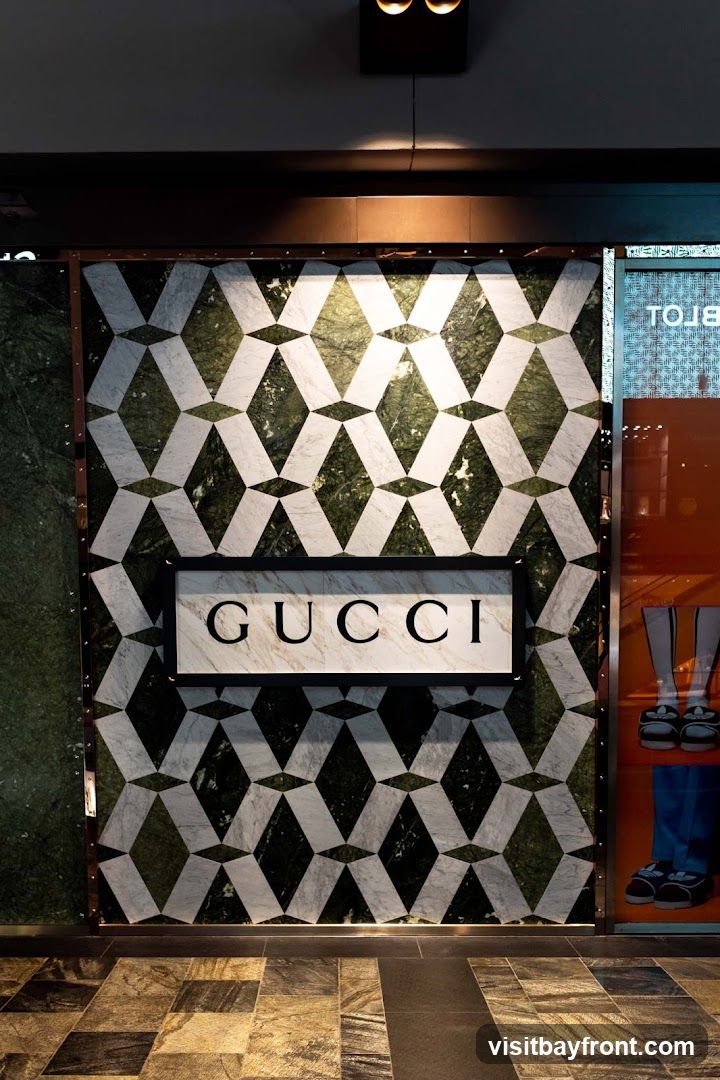 A Gucci store facade featuring a geometric marble pattern in green and white. A rectangular sign with the word "GUCCI" in black lettering is centered on the facade. To the right, a display window shows mannequins wearing shoes and boots. The floor is tiled with a mosaic of brown and grey stones.