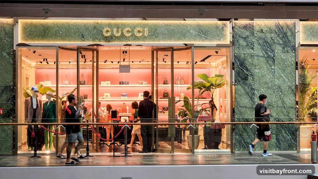 The exterior of a Gucci store with mannequins displaying clothing and accessories in the window. The store features a marble facade and a prominent "GUCCI" sign above the entrance. People are seen entering and walking past the store.