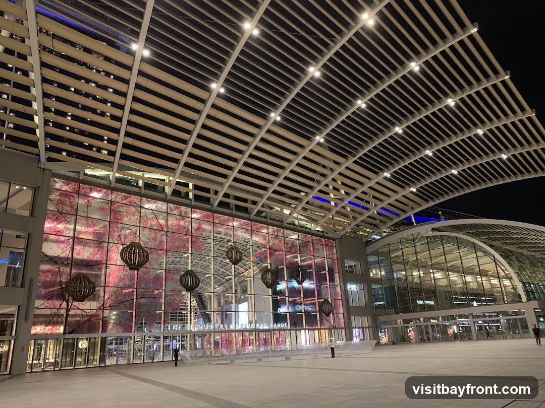 A wide shot of the exterior of The Shoppes at Marina Bay Sands at night. The building features a large, curved glass facade with a modern, slatted roof structure overhead. The glass wall is illuminated with a vibrant pink cherry blossom pattern. Several large, spherical lanterns hang in front of the glass. The entrance to the mall is visible, with people walking in and out.