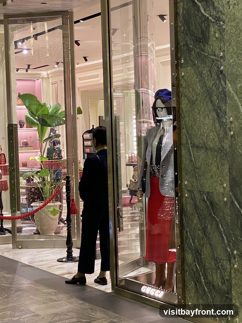 A view of a luxury store's entrance, featuring a mannequin dressed in a sparkly blazer, white shirt, black tie, and a red skirt. The store interior is visible, showcasing handbags on display shelves and a large potted plant. The store's name,