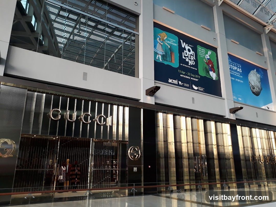 The exterior of a Gucci store with its prominent logo above the entrance. The facade is made of reflective, vertical panels. Inside, mannequins display clothing. Above the store, large posters advertise exhibitions for ACMI and ArtScience Museum.