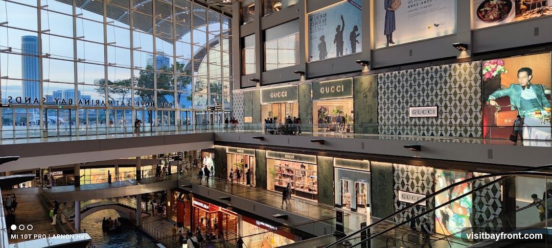 The image shows the interior of a multi-level shopping mall. Several Gucci stores are visible with their distinctive signage. The mall features large glass windows offering a view of the city skyline and a canal with boats. There are also advertisements and displays on the walls. People are seen shopping and walking around.
