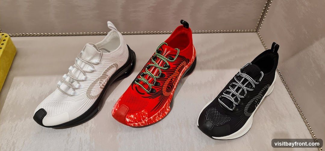 Three athletic shoes are displayed on a textured surface. From left to right, the shoes are white, red, and black. The white shoe has a subtle grey logo on the side. The red shoe features a black and orange graphic design and bright green laces. The black shoe has a grey logo on the side and white and black laces. A yellow object with holes is visible on the far left. The display is framed by a decorative silver beaded trim.