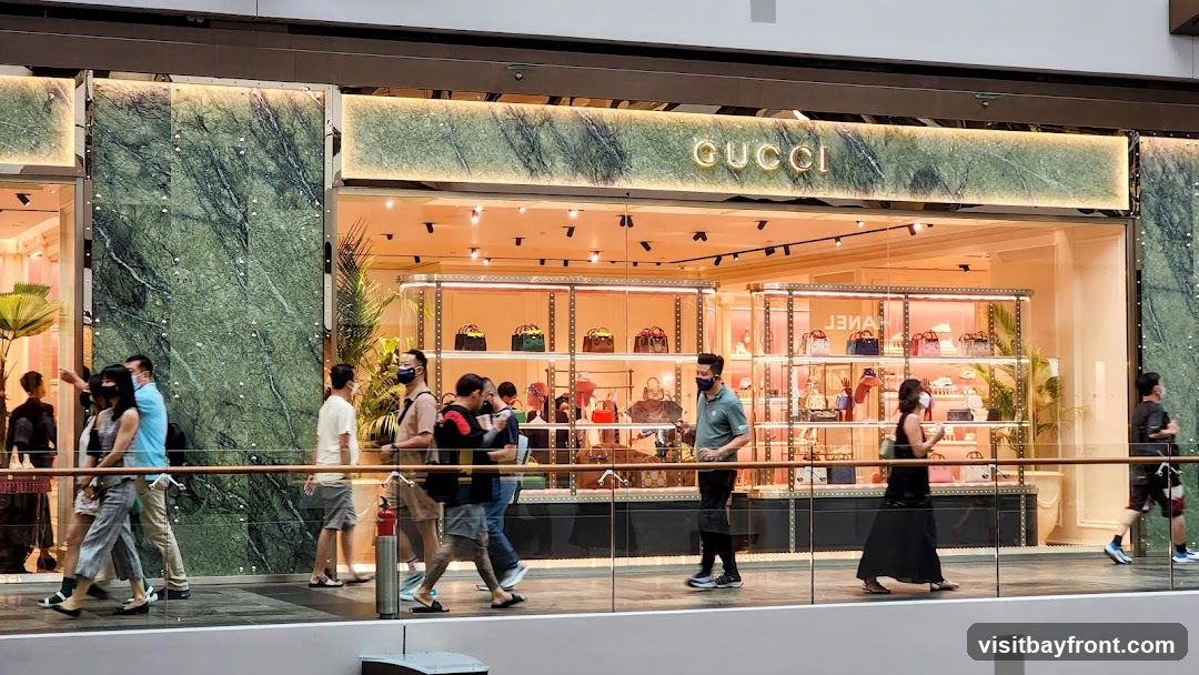 A Gucci store with a marble facade and illuminated signage. The interior displays various handbags and accessories on shelves. Several people are walking past the storefront.