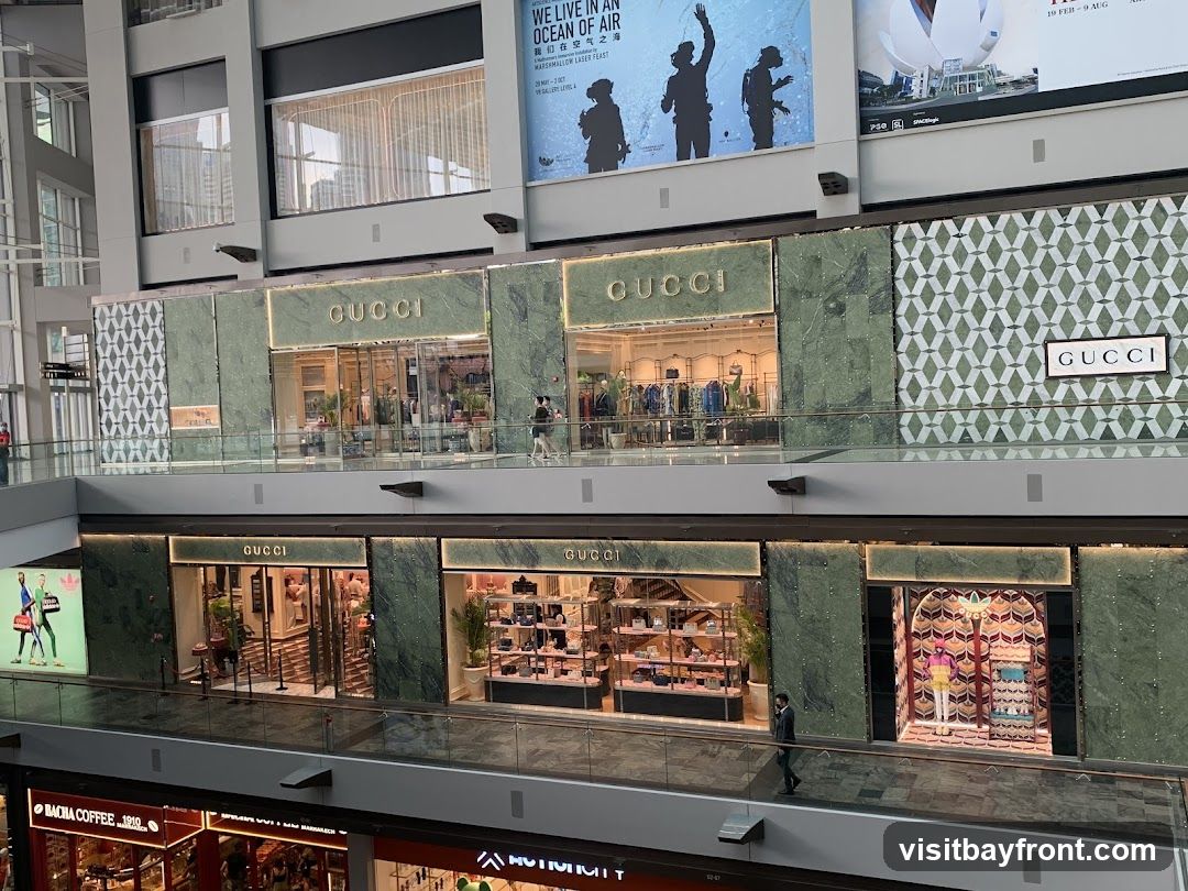 Multiple Gucci storefronts are visible on two levels of a shopping mall. The stores feature green marble facades and large display windows showcasing merchandise. Above the upper level stores, large advertisements are displayed.