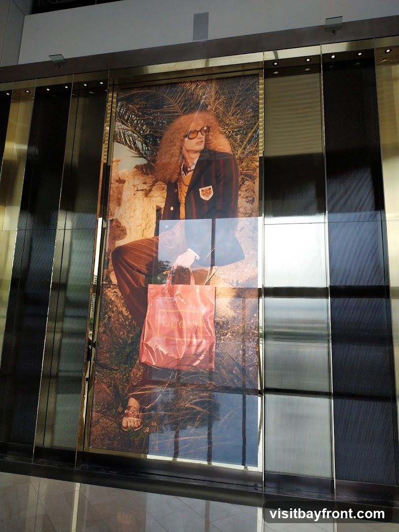 A large advertisement featuring a person with voluminous curly hair wearing sunglasses, a striped blazer with a crest, and brown pants. The person is holding a red Gucci shopping bag. The advertisement is displayed on a building facade with reflective panels.