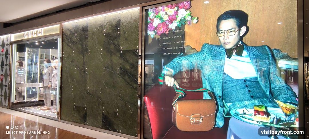 A large display features a man in a plaid suit, holding a Gucci handbag. He is seated next to a table with a dessert and a cocktail. To the left, the entrance to a Gucci store is visible, with mannequins dressed in suits displayed in the window. The store's name, "GUCCI," is illuminated above the entrance.