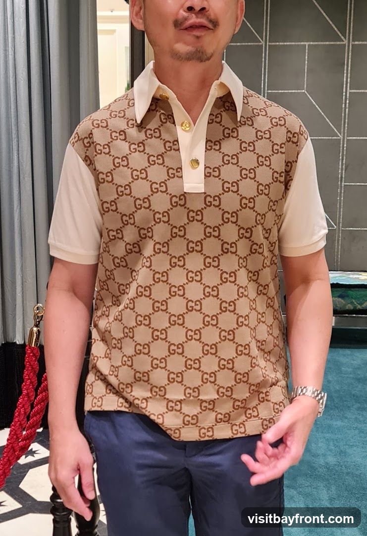 A man is shown from the chest down, wearing a short-sleeved polo shirt. The shirt features a repeating pattern of the Gucci logo in brown on a beige background, with cream-colored sleeves and collar. The collar has two gold buttons. The man is also wearing dark blue trousers and a silver watch on his left wrist. The background appears to be an indoor setting, possibly a store, with a red velvet rope barrier in the foreground and a patterned wall behind.