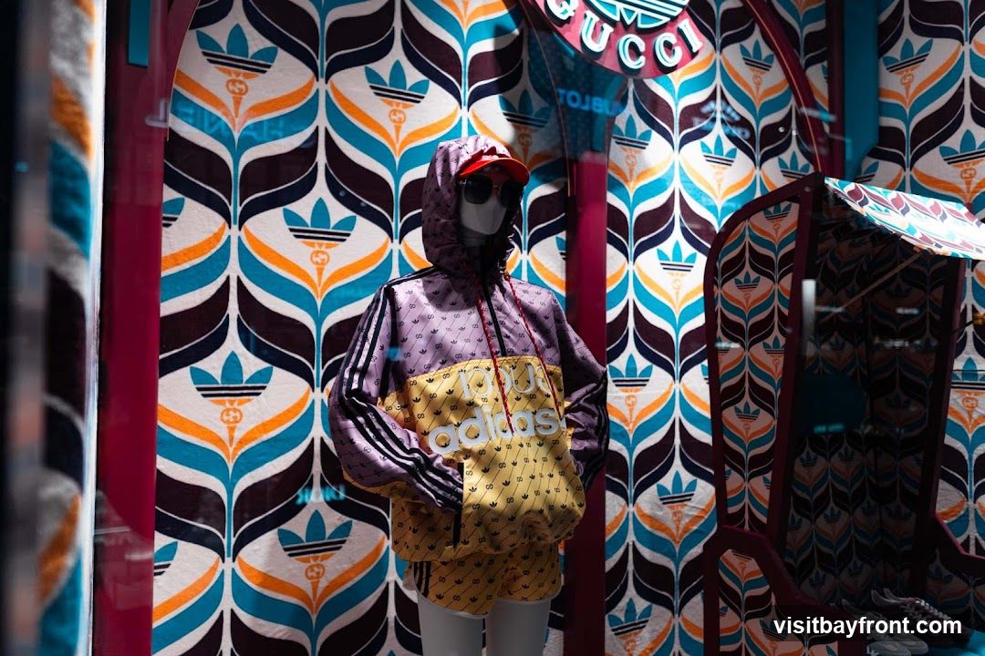 A mannequin is dressed in a purple and yellow Adidas x Gucci tracksuit, complete with a hooded jacket and shorts. The mannequin wears sunglasses and a red cap. The background features a vibrant, repeating pattern of stylized leaves and flowers in shades of blue, orange, brown, and white, incorporating both the Adidas trefoil logo and the Gucci logo. To the right, a display structure echoes the patterned background, and a pair of Adidas sneakers are visible.