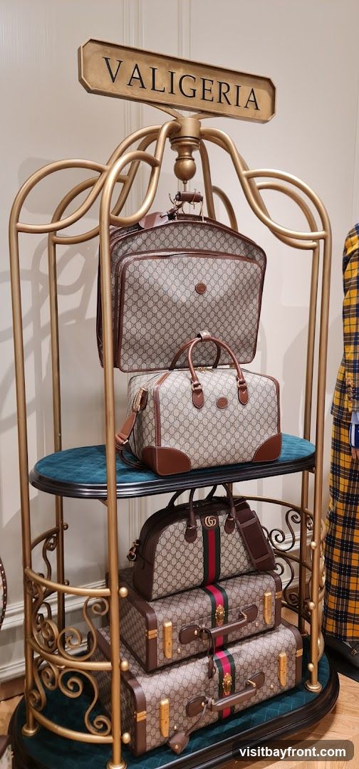 A display of stacked luggage, featuring the Gucci monogram pattern and brown leather trim. The luggage is presented on a gold-colored stand with decorative scrollwork. A sign above reads "VALIGERIA". A person in a yellow and blue plaid jacket is partially visible on the right.