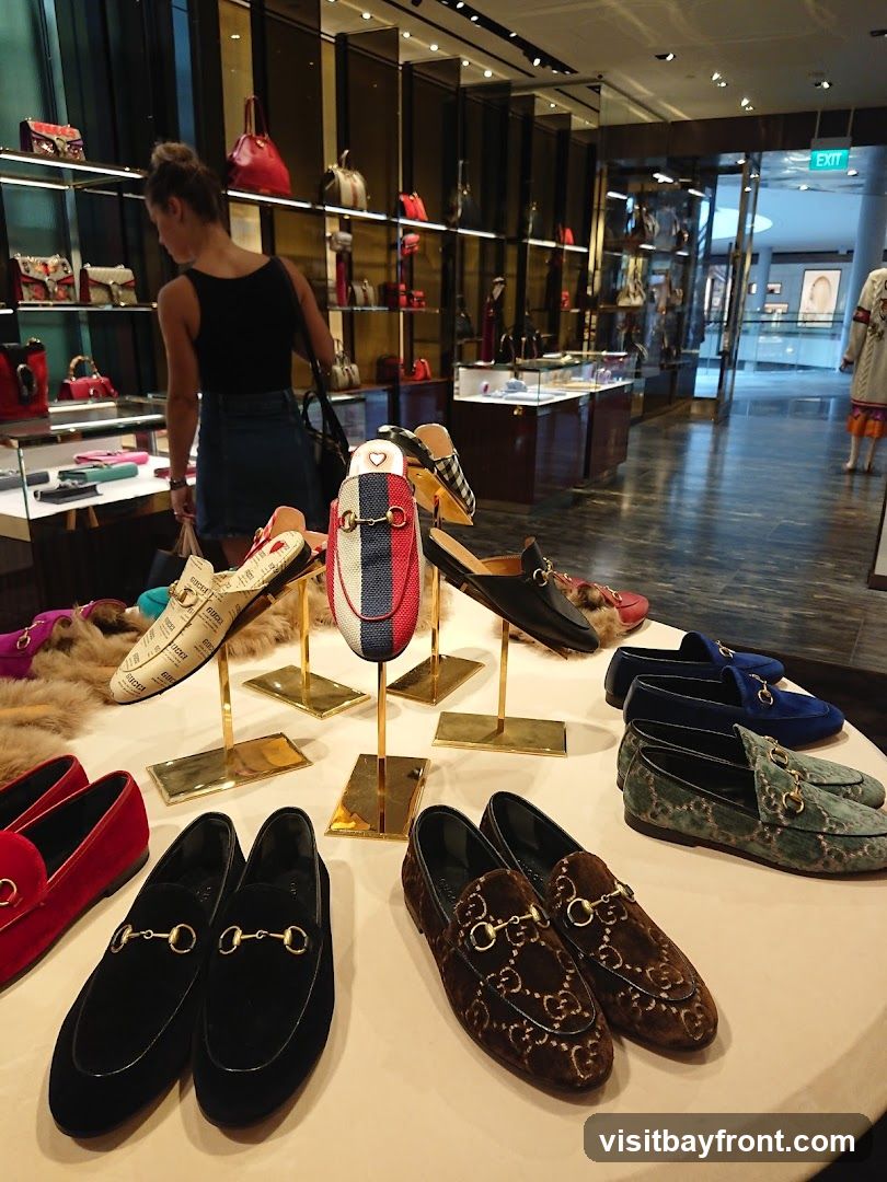 A display of various luxury loafers and mules are showcased on a table in a well-lit boutique. In the background, a woman browses handbags on shelves, and the store interior with glass doors and displays is visible.