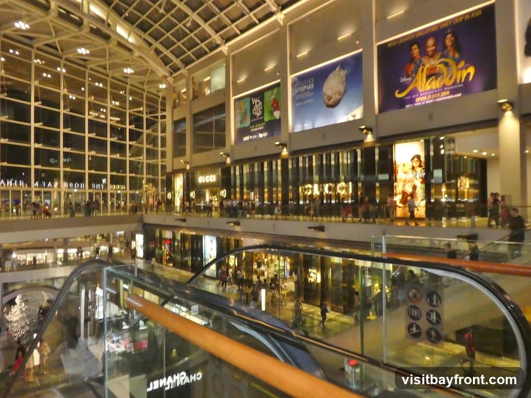 The image shows the interior of a large, modern shopping mall with multiple levels. Escalators are visible, along with glass railings and a reflective floor. Several shops are visible, including Gucci. Large advertisements are displayed on the upper walls, including one for Disney's Aladdin musical. People are seen browsing and walking around the mall.