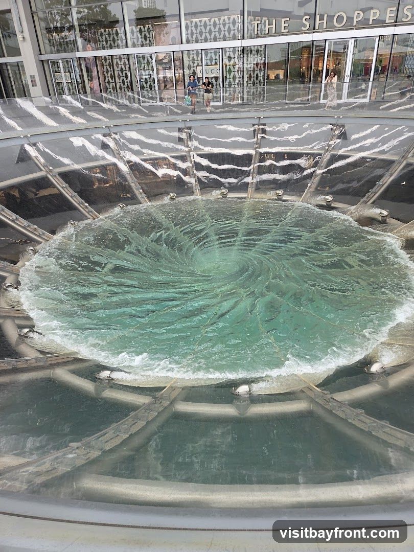 A large, circular fountain with swirling turquoise water is in the foreground. In the background, the glass facade of a building with the words "THE SHOPPES" is visible, along with several storefronts, including one with a "GUCCI" sign.