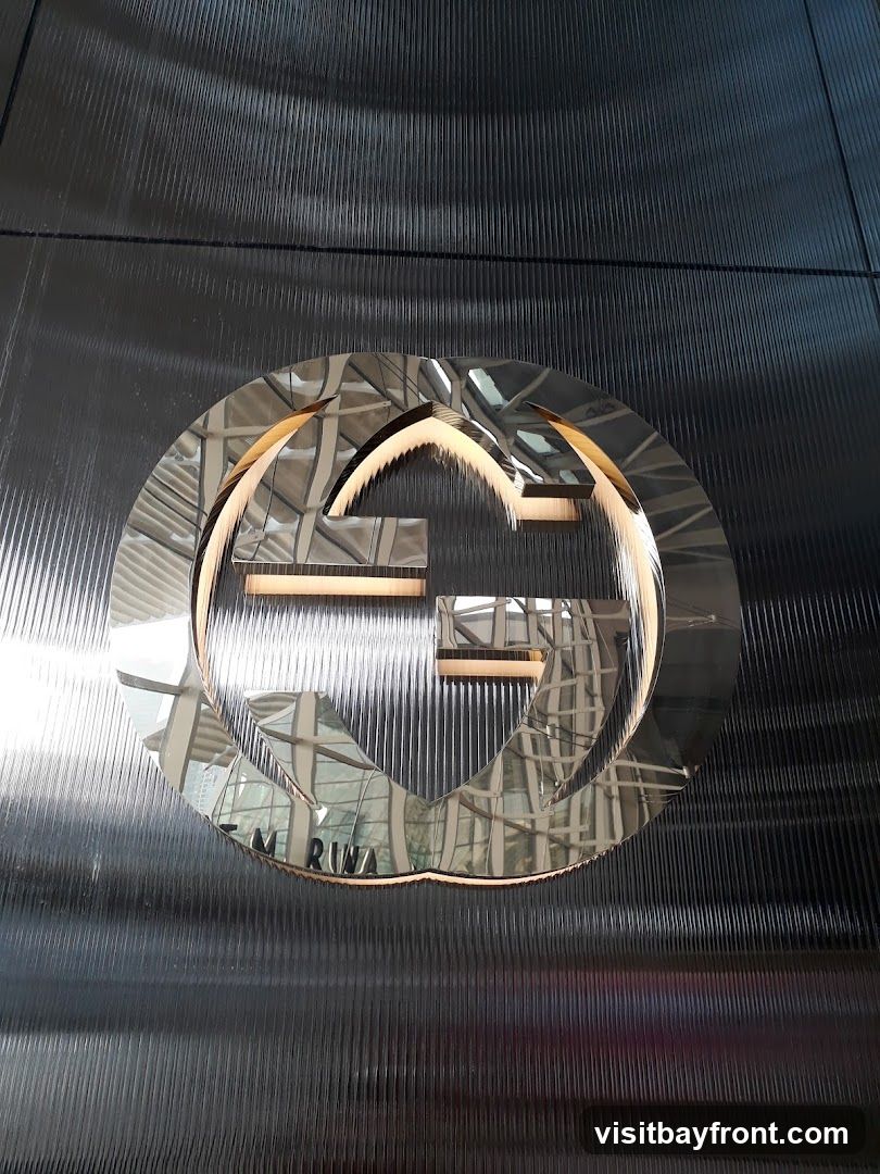 A close-up shot of a large, metallic Gucci logo mounted on a dark, ribbed wall. The logo is a double G symbol within a circle, with a polished, reflective surface that mirrors the interior of a building, showing architectural details and lighting. Below the logo, partially obscured text reads "M RUWA" which appears to be part of a larger sign or inscription.