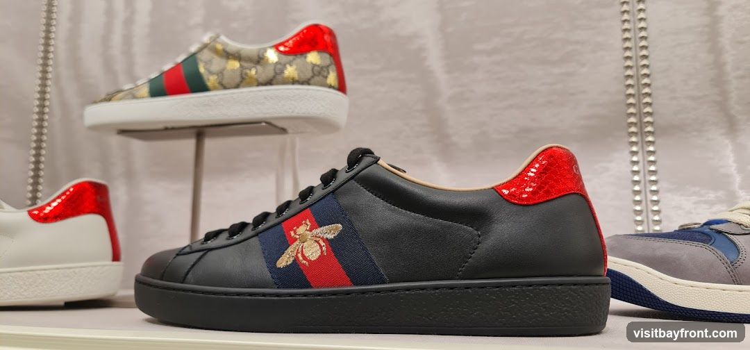 A close-up shot of several pairs of designer sneakers displayed on a shelf. The focus is on a black leather sneaker with red accents and a bee emblem on a striped band. Another sneaker with a gold and beige monogram pattern and red accents is visible in the background, along with a partial view of a white sneaker and a blue and grey sneaker.