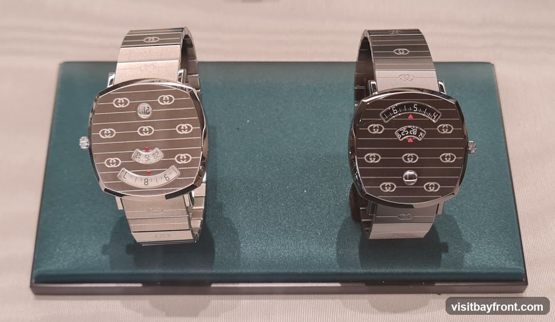 Two silver Gucci watches are displayed on a teal stand. The watch on the left shows the date as 12, and the time as approximately 8:32. The watch on the right shows the time as approximately 5:25-29. Both watches have the Gucci logo repeated across the face and on the bands.