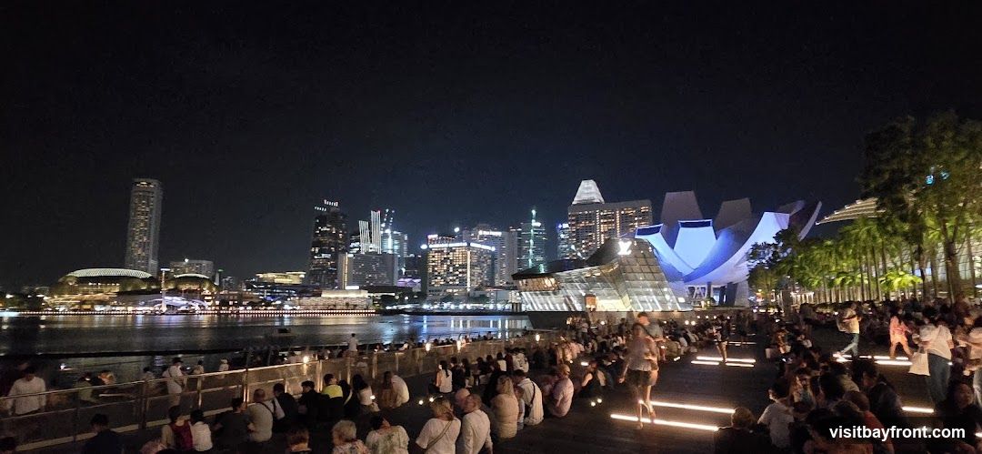 A panoramic night view of a city skyline across a body of water. The skyline features numerous illuminated skyscrapers and distinctive modern architecture, including a building with a lotus-like structure. In the foreground, a large crowd of people are seated and standing on a wooden promenade, facing the water and the city. The promenade is lit with linear lights embedded in the ground. Palm trees are visible on the right side of the image.