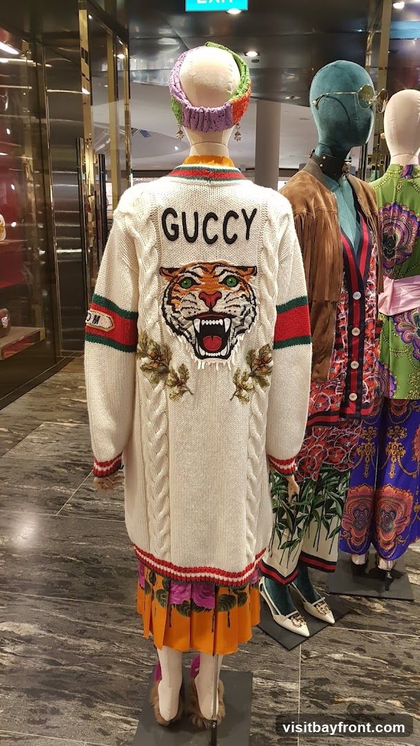 A display of mannequins wearing fashionable clothing. The central mannequin wears a cream-colored cable-knit sweater with a tiger face and the word "GUCCY" embroidered on the back. It is paired with a pleated orange skirt with a floral print. Other mannequins are dressed in colorful, patterned garments. The background shows a store interior with reflective surfaces and an exit sign.
