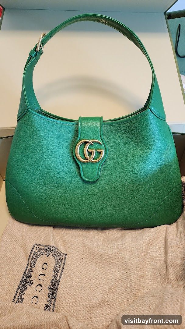 A green leather Gucci shoulder bag with a gold GG logo clasp is displayed. The bag is resting on a beige dust bag with a decorative Gucci logo printed on it.