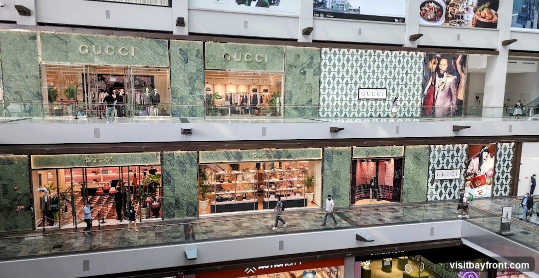 Multiple Gucci storefronts are visible on two levels of a shopping mall. The facades feature green marble accents and large glass windows displaying merchandise. Some storefronts have large advertisements or patterned wallpaper. People are seen walking on the elevated walkways in front of the stores.