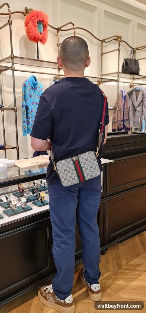 A man stands in a store, facing away from the camera. He is wearing a dark blue t-shirt and blue pants. He has a Gucci crossbody bag with the signature GG pattern and green and red stripe. The bag is worn across his body. In the background, there are shelves displaying clothing and accessories, including a bright blue sweater with a colorful pattern, a grey blazer, and a black handbag. A display case in the foreground shows several watches. The man is also wearing Gucci sneakers.