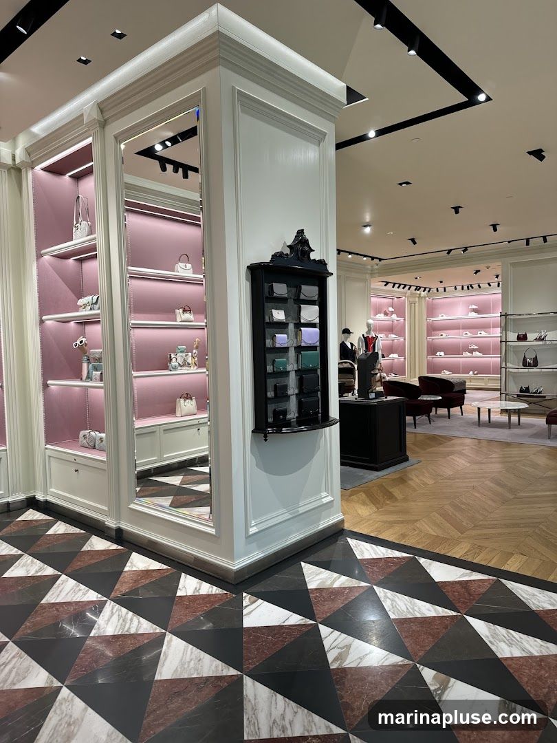 The interior of a high-end retail store, likely a fashion boutique. The foreground features a striking geometric floor pattern in marble and dark stone. To the left, illuminated shelves display various handbags and accessories against a pink backdrop. A black display cabinet with multiple compartments holding small leather goods is mounted on a white pillar. In the background, more display shelves with merchandise are visible, along with seating areas and mannequins dressed in clothing. The overall ambiance is elegant and sophisticated, with clean lines and soft lighting.