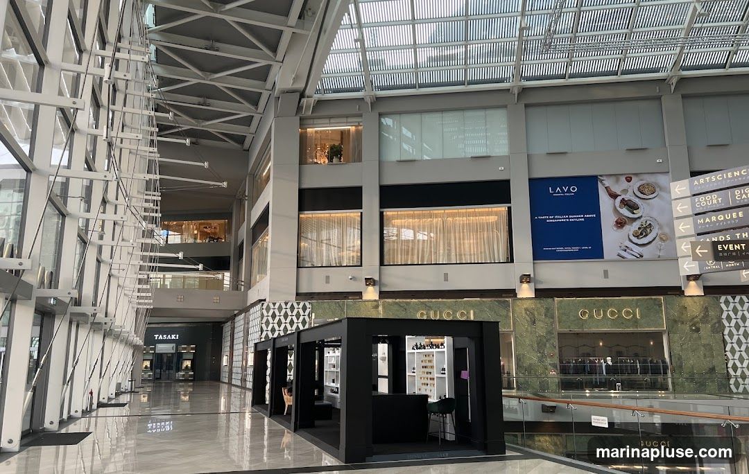The image shows the interior of a spacious, modern shopping mall with a high ceiling and a glass roof. On the left, a large glass facade with a white metal structure runs along the walkway. In the center, there are several shops. A prominent Gucci store with a green marble facade and gold lettering is visible. In front of it, a black, modern kiosk displays various items. Further down the walkway, a store with the name TASAKI is visible. On the right, directional signs point to different areas like