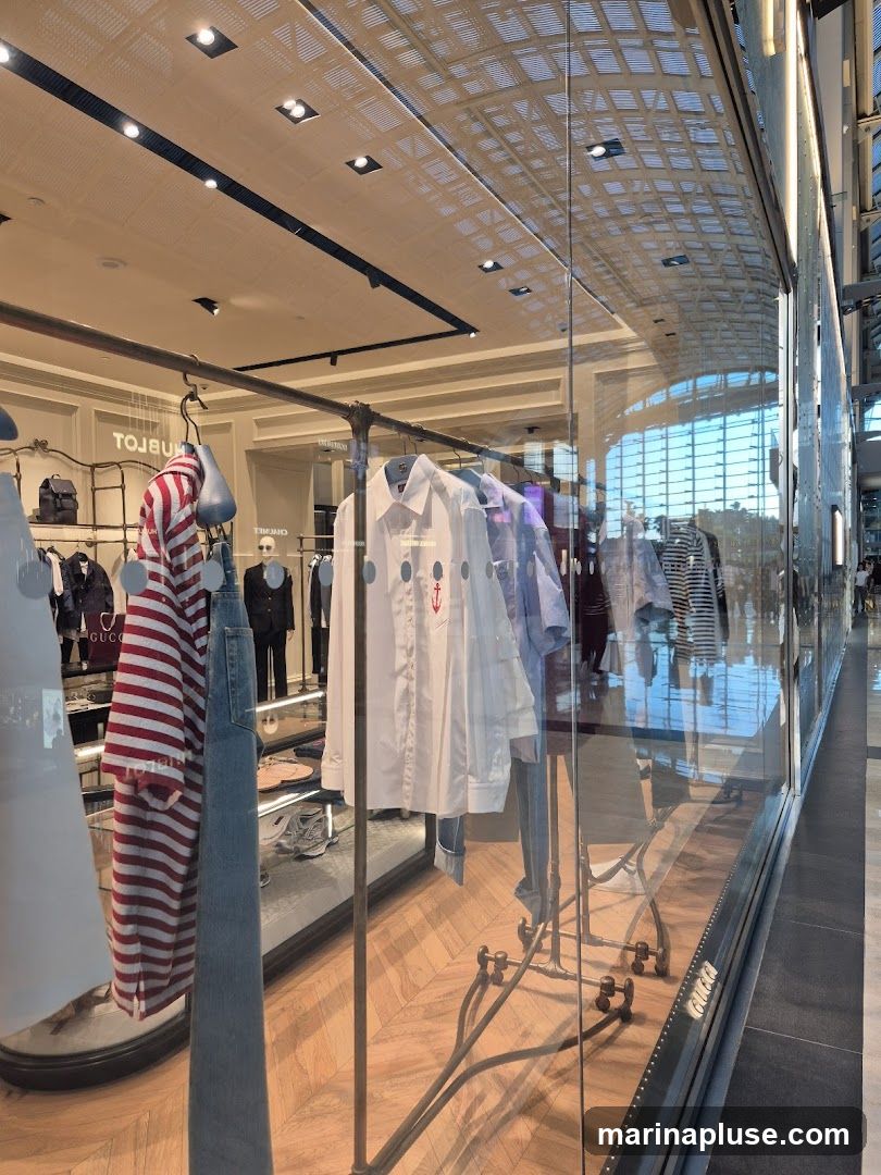 A view through a glass window into a clothing store. Racks of clothing, including a red and white striped shirt, white shirts with circular patterns, and blue jeans, are displayed. Mannequins are also visible inside the store, along with shelves of accessories. The store interior features a wooden floor and a patterned ceiling with lighting. Reflections of the outside environment are visible on the glass.