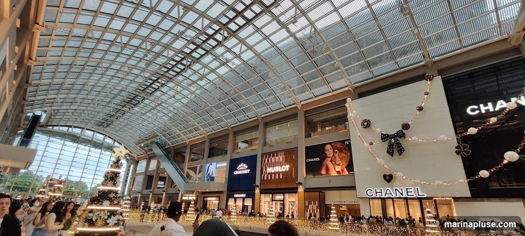 An interior view of a large, modern shopping mall with a distinctive glass roof. Several luxury brand stores are visible, including Chanel, Hublot, and Chaumet, with their prominent signage. The mall is decorated for the holidays with Christmas trees and festive lights, and many shoppers are present.