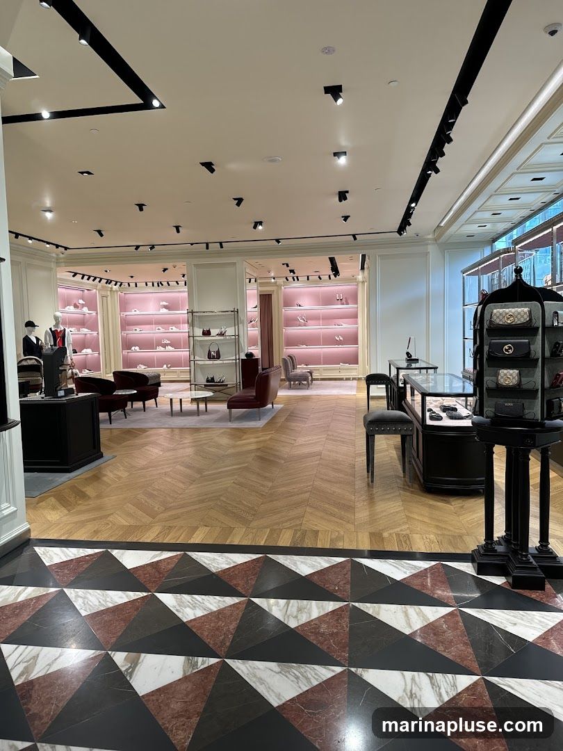 The interior of a high-end retail store, likely a fashion boutique, is shown. The space features polished wooden floors with a geometric marble inlay at the entrance. The walls are lined with display shelves illuminated by soft pink lighting, showcasing an array of shoes and handbags. Mannequins dressed in formal attire are positioned near the entrance. Seating areas with plush armchairs and sofas are arranged throughout the store. Display cases with glass tops exhibit accessories. The overall ambiance is elegant and sophisticated, with modern track lighting illuminating the merchandise.