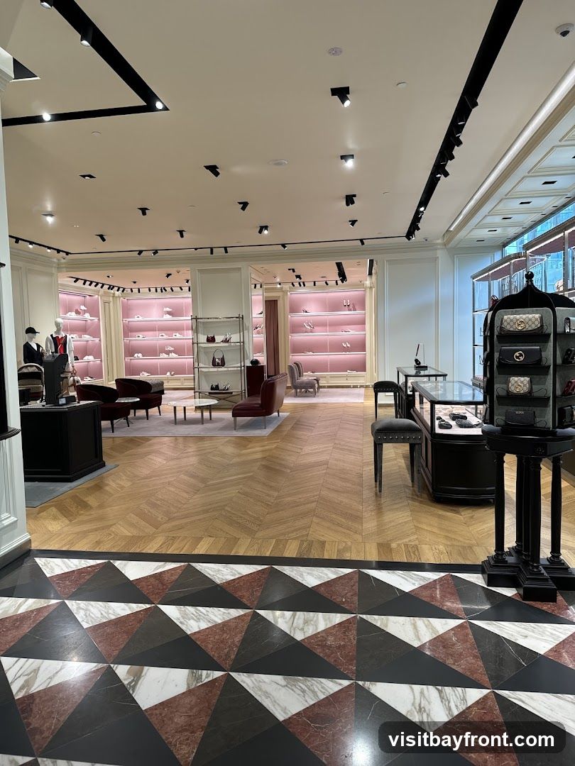 The interior of a high-end retail store, likely a fashion boutique, is shown. The space features polished wooden floors with a geometric marble inlay at the entrance. The walls are lined with display shelves illuminated by soft pink lighting, showcasing an array of shoes and handbags. Mannequins dressed in formal attire are positioned near the entrance. Seating areas with plush armchairs and sofas are arranged throughout the store. Display cases with glass tops exhibit accessories. The overall ambiance is elegant and sophisticated, with modern track lighting illuminating the merchandise.