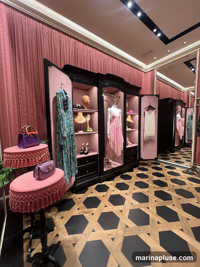 A well-lit boutique interior with plush pink velvet curtains and display cabinets. The cabinets showcase various high-end fashion items, including dresses on mannequins, handbags, jewelry, and shoes. The floor features a distinctive geometric pattern of wood and black tiles. Mirrors reflect the displays, creating a sense of depth.
