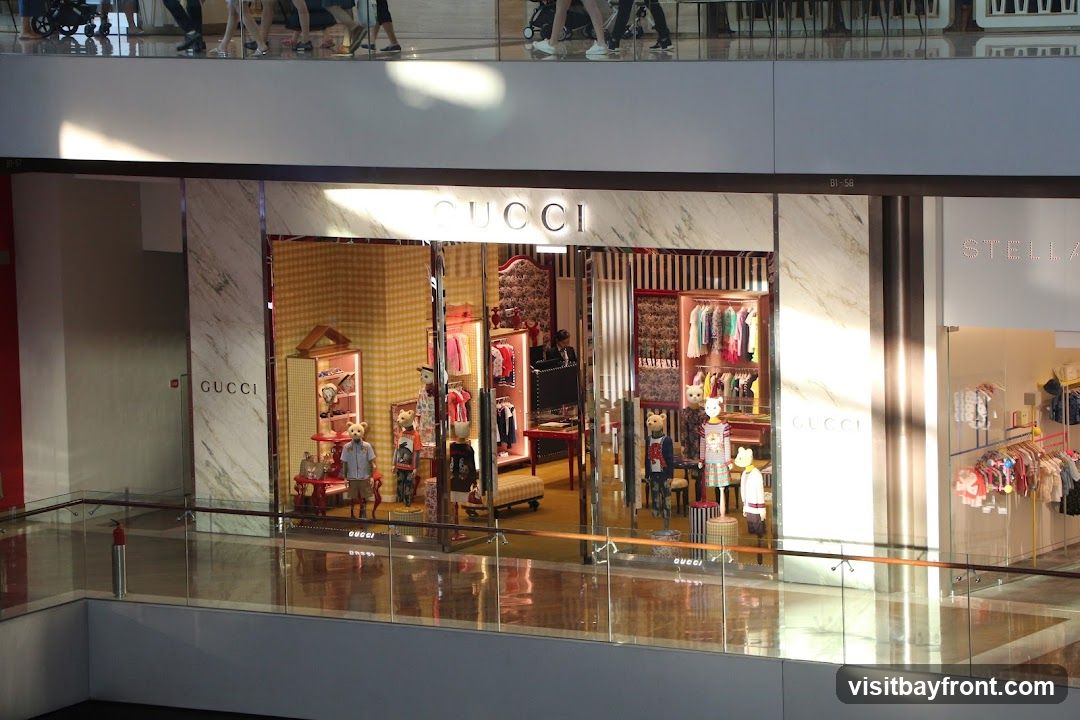 The exterior of a Gucci store with a large display window. The store features mannequins dressed in children's clothing and teddy bears. The Gucci logo is visible on the marble facade and on the glass doors.