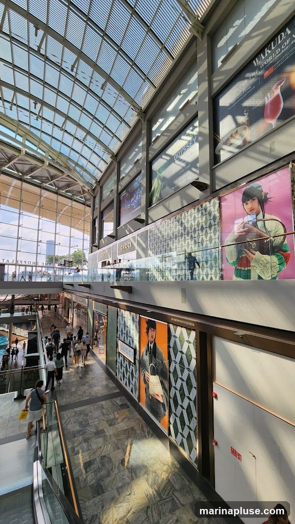 A wide-angle shot of the interior of a modern shopping mall, featuring a glass ceiling that allows natural light to filter in. On the upper level, large advertisements are displayed, including one for "Wakuda The World of..." and another for "Odyssey". Below, the Gucci store is visible with its distinctive logo and patterned facade. Several shoppers are seen walking along the elevated walkway and on the lower level. The overall impression is one of a bustling, upscale retail environment.