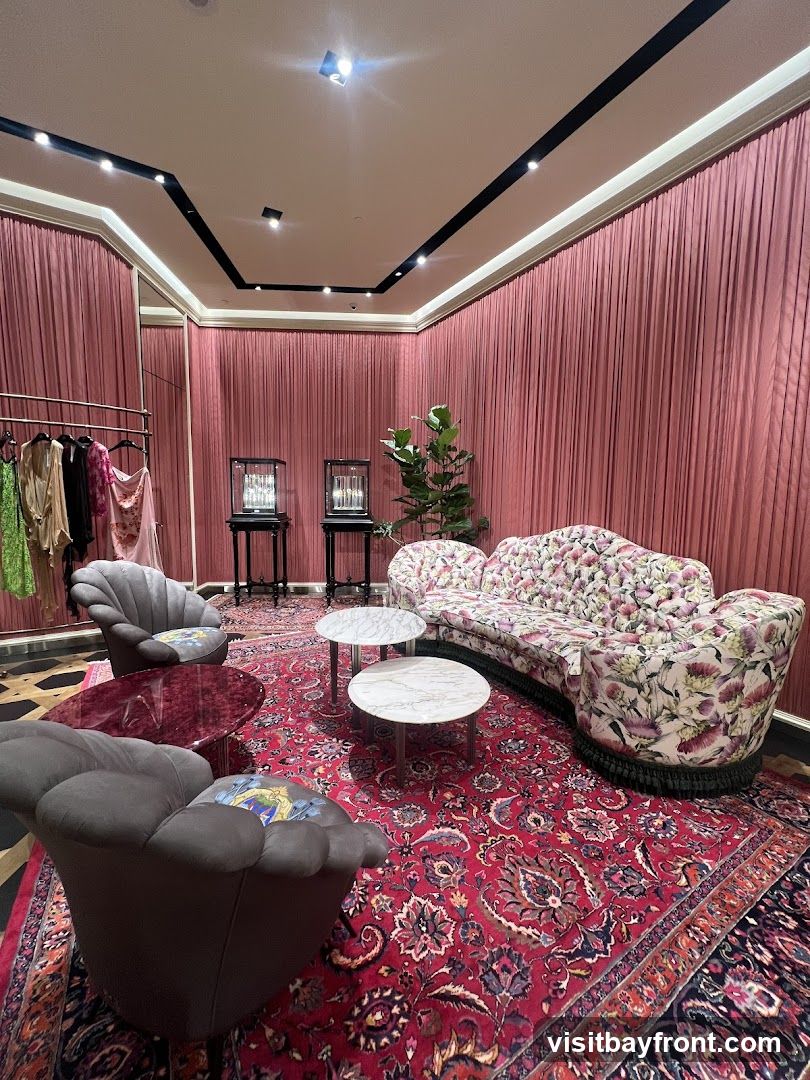 A plush lounge area with pink velvet curtains, a floral-patterned sofa, two grey armchairs, and a red patterned rug. Several small coffee tables are placed around the seating. In the background, there is a clothing rack with garments and two display cases. A potted plant adds a touch of greenery.