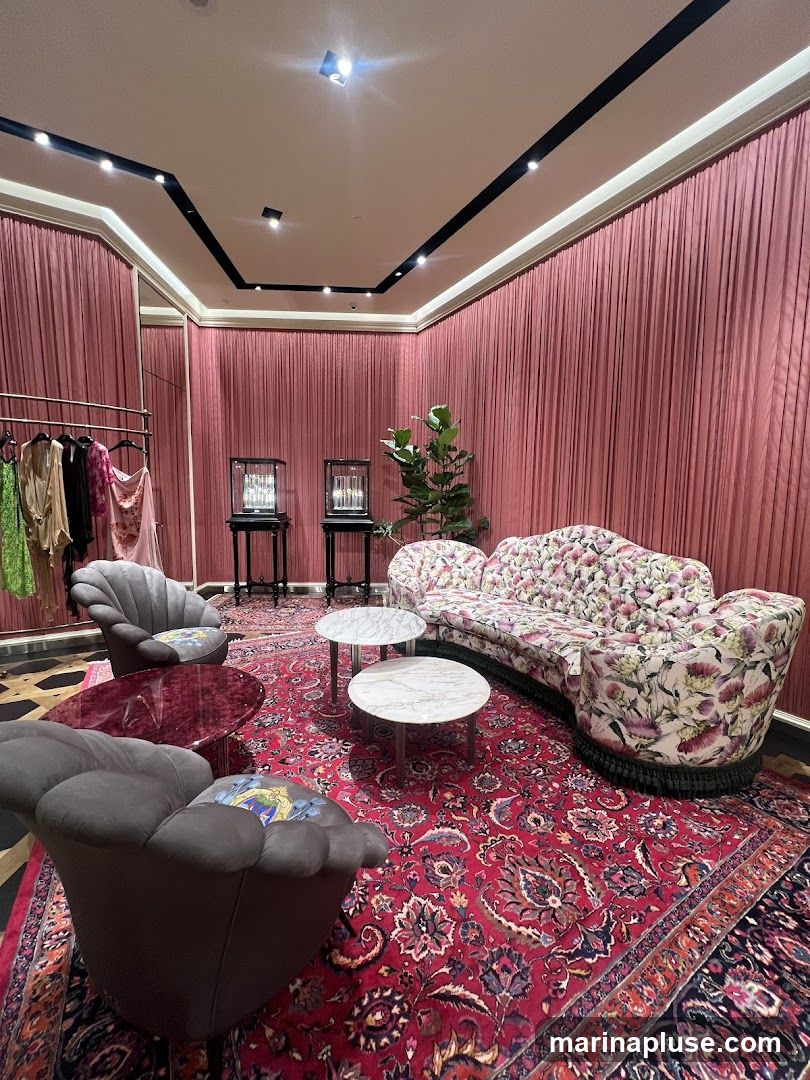 A plush lounge area with pink velvet curtains, a floral-patterned sofa, two grey armchairs, and a red patterned rug. Several small coffee tables are placed around the seating. In the background, there is a clothing rack with garments and two display cases. A potted plant adds a touch of greenery.