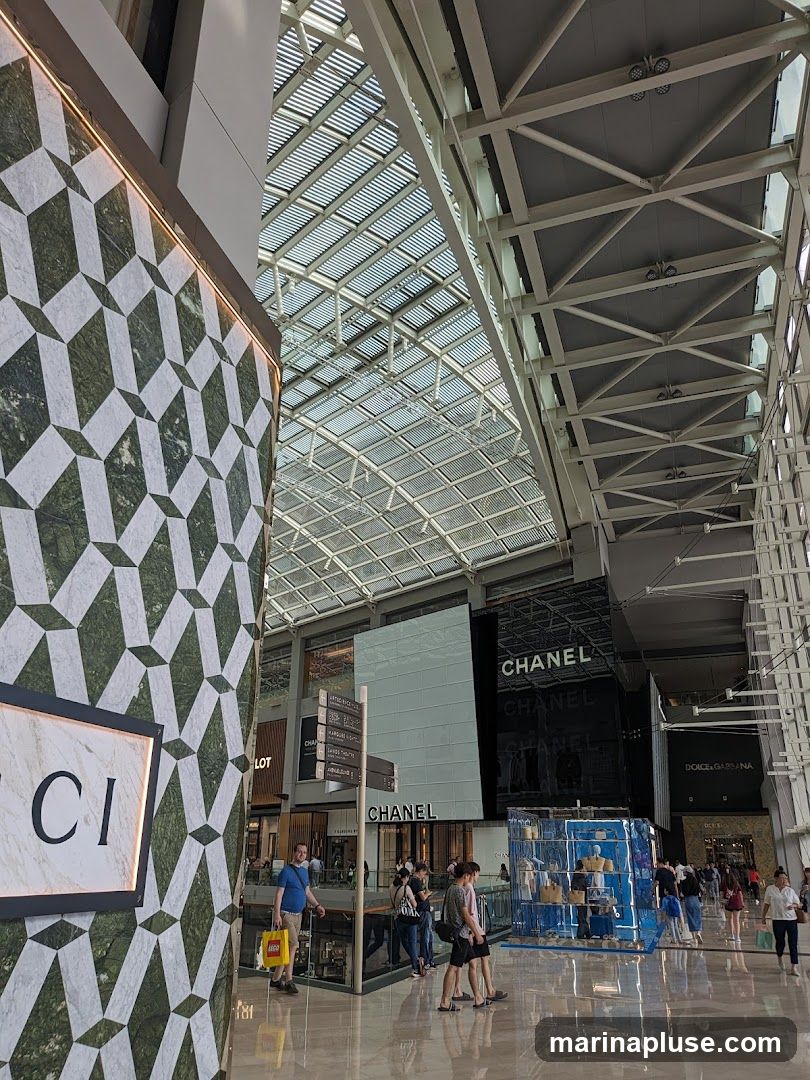 An interior view of a modern shopping mall with a high, glass-paneled ceiling and exposed metal beams. On the left, a section of a Gucci store facade is visible, featuring a distinctive green and white geometric pattern and a sign with the brand's logo. Further into the mall, large signs for Chanel and Dolce & Gabbana are prominent. Several people are walking through the mall, and mannequins are displayed in a blue-lit booth.