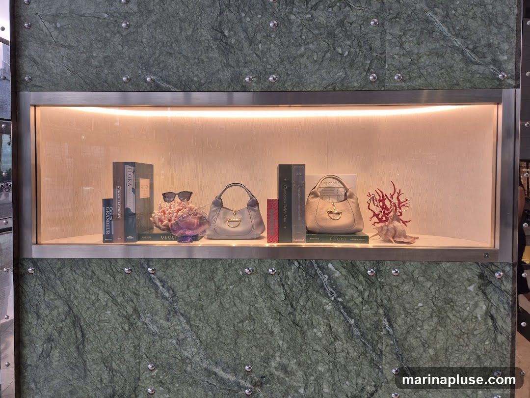A display window features two light-colored Gucci handbags. The handbags are placed on a shelf with decorative items including books, sunglasses, and coral. The background of the display is illuminated with warm lighting. The exterior of the display window is framed by green marble-like panels with silver accents.