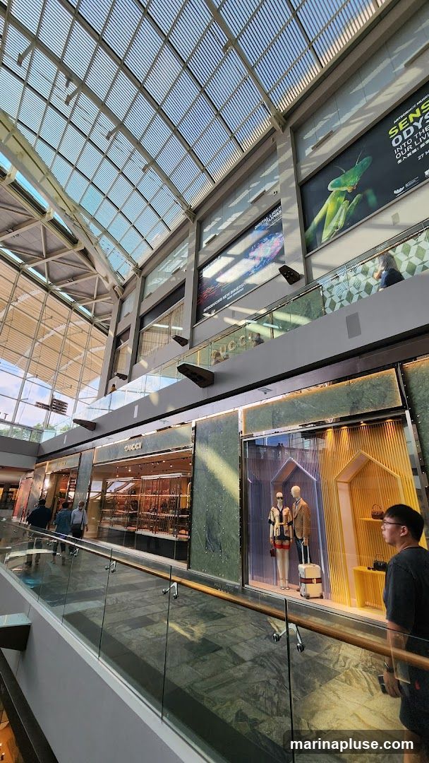 A wide-angle shot of the interior of a modern shopping mall, looking upwards towards a large, slatted glass ceiling. On the lower level, a Gucci store is visible with its name displayed prominently. Adjacent to it is a display window featuring mannequins dressed in fashionable clothing and a suitcase. On the upper levels, large digital screens display advertisements, including one for an art exhibition with a praying mantis image. People are seen walking along the elevated walkways and browsing the shops. The architecture features sleek lines, marble accents, and glass railings.