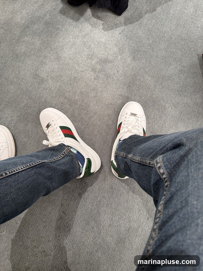 A top-down view of a person wearing white sneakers with green and red stripes on a grey carpeted floor. The person is also wearing blue jeans.