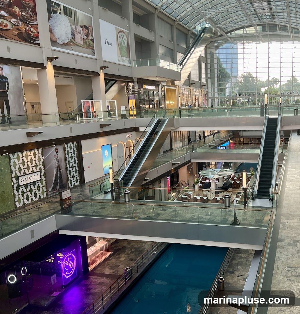 The image shows the interior of a multi-level shopping mall with escalators, shops, and a canal. Large advertisements for brands like Dior and Gucci are visible on the walls. People are seen walking on the different levels. The mall has a glass ceiling, allowing natural light to enter.