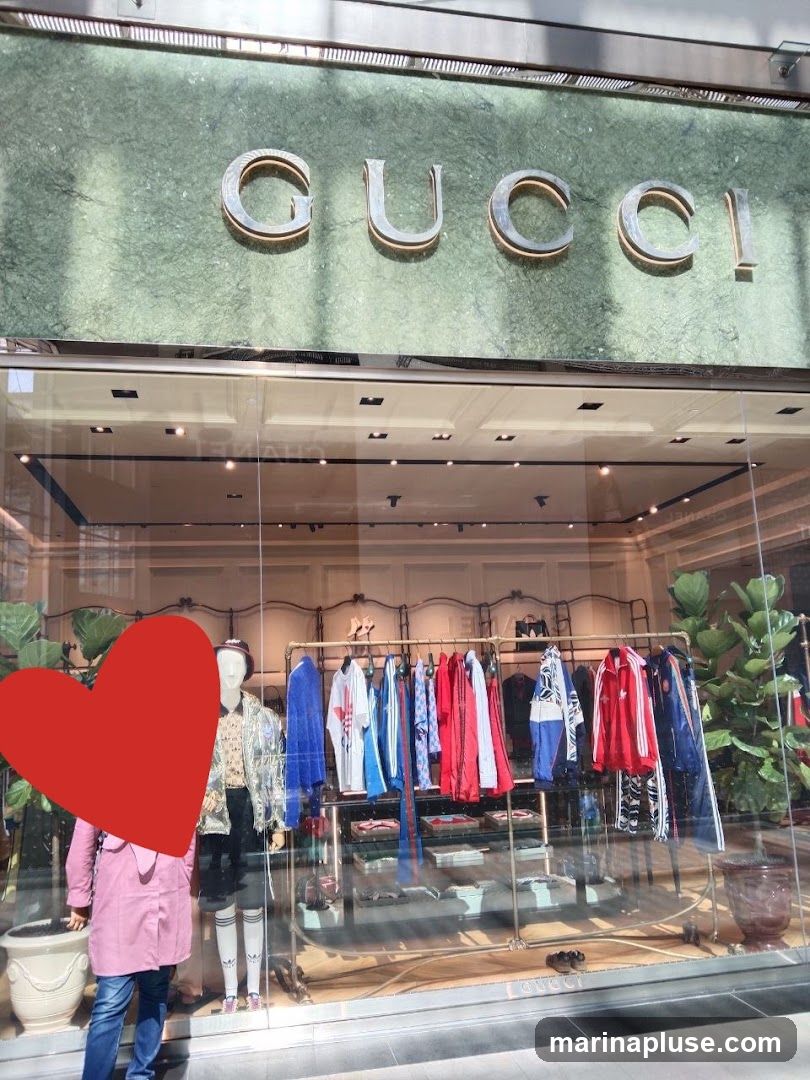 The exterior of a Gucci store with the brand name prominently displayed in large metallic letters above the entrance. The storefront features a large display window showcasing clothing on racks and mannequins. Lush green plants are visible on either side of the display. A red heart shape obscures part of a mannequin in the foreground.