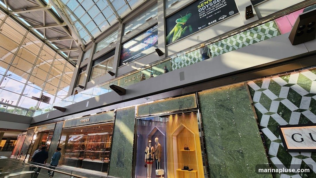 A view of a Gucci store interior and exterior, with mannequins displaying clothing and accessories. The store features marble paneling and a large glass facade. Above the store, there are advertisements for an exhibition.