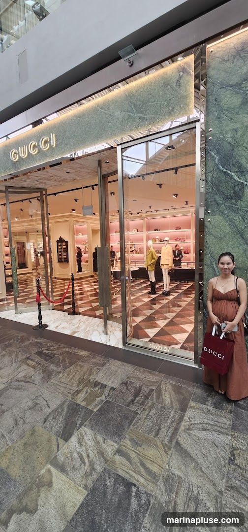 A woman stands to the right of a Gucci store entrance. The store has a large glass facade with the Gucci logo visible above the entrance. Inside, mannequins are displayed, and shelves are stocked with merchandise. The floor inside and outside the store is tiled.