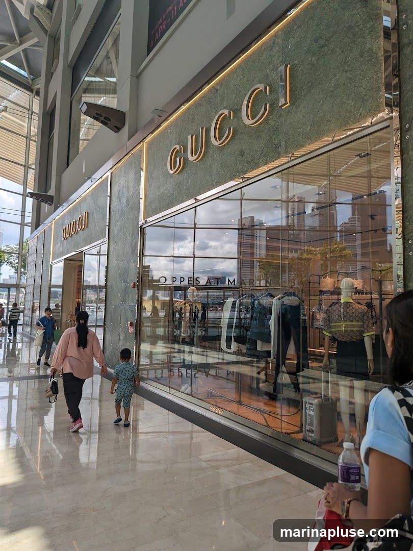The exterior of a Gucci store with large glass windows displaying clothing and mannequins. The Gucci logo is prominently displayed in gold lettering on a green marble facade above the windows. People are walking by the storefront in a well-lit indoor walkway.