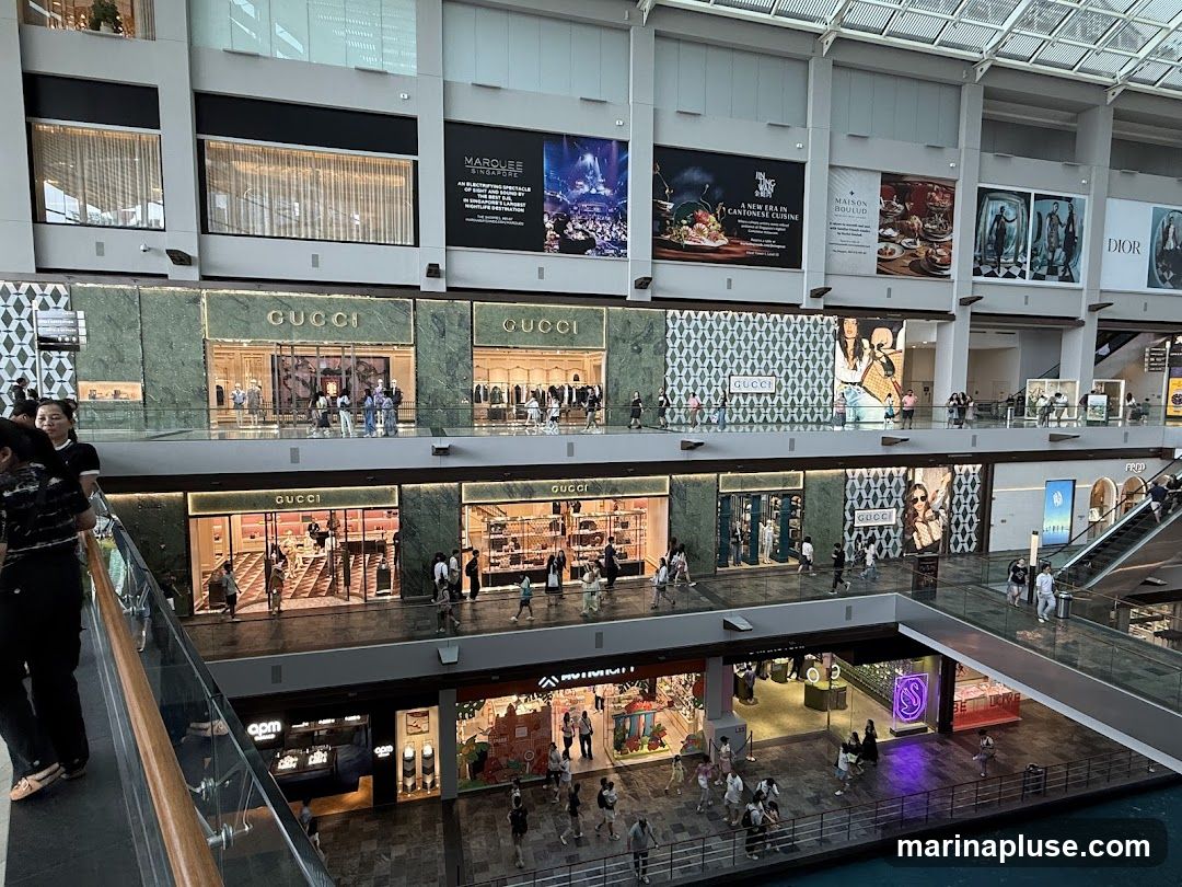 The image shows the interior of a large, modern shopping mall with multiple levels. Several storefronts are visible, including multiple Gucci stores with their distinctive green marble facade and gold lettering. Large advertisements and banners are displayed on the upper levels, featuring brands like Dior and Marquee Singapore. People are seen walking around, browsing in stores, and using escalators. The overall impression is a busy and upscale retail environment.