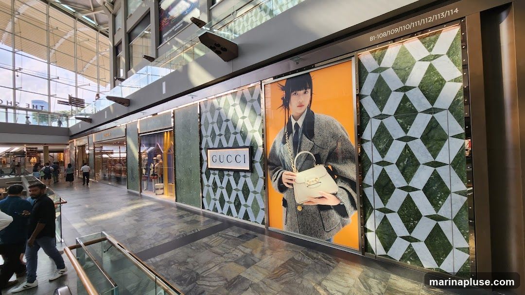 A wide shot of a Gucci store's exterior display in a modern shopping mall. The display features a large advertisement of a woman holding a handbag, flanked by patterned panels. The Gucci logo is prominently displayed on a white rectangular sign. Other storefronts and shoppers are visible in the background.