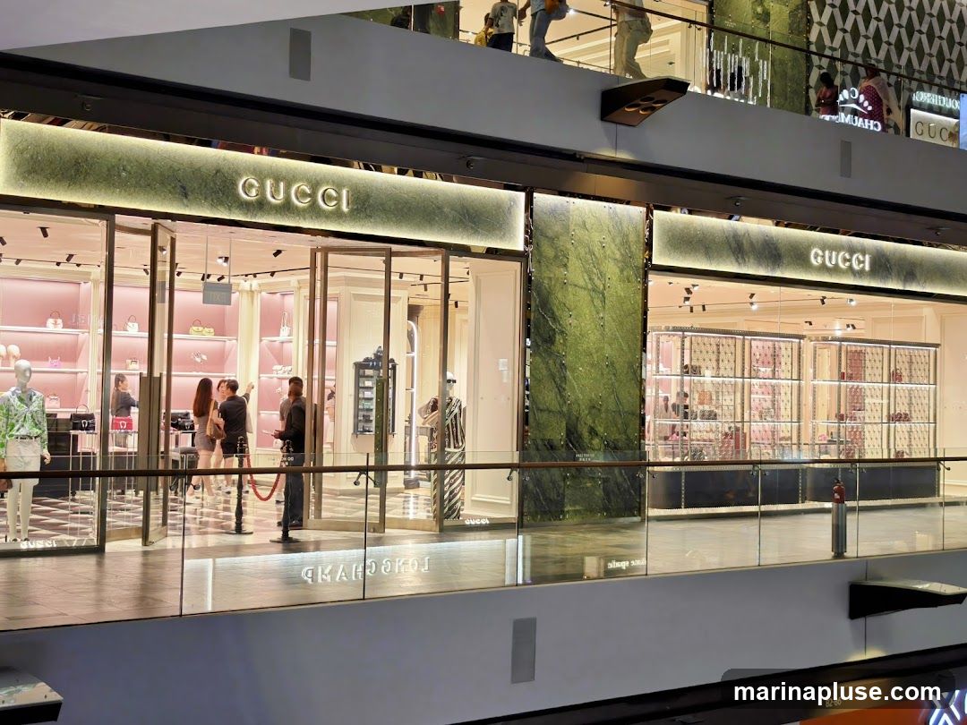 The exterior of a Gucci store in a mall. The store has a large glass facade with the Gucci logo prominently displayed above the entrance. Inside, shelves are stocked with handbags and accessories, illuminated by soft lighting. Mannequins display clothing.