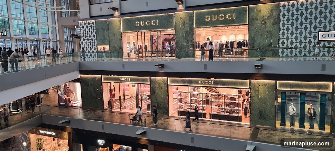 A wide shot of a multi-level shopping mall interior, focusing on two levels of Gucci storefronts. The upper level features two distinct Gucci entrances with illuminated signage and displays of clothing. The lower level also shows multiple Gucci entrances, with displays of handbags and accessories in glass cases. People are seen walking on the elevated walkways in front of the stores.