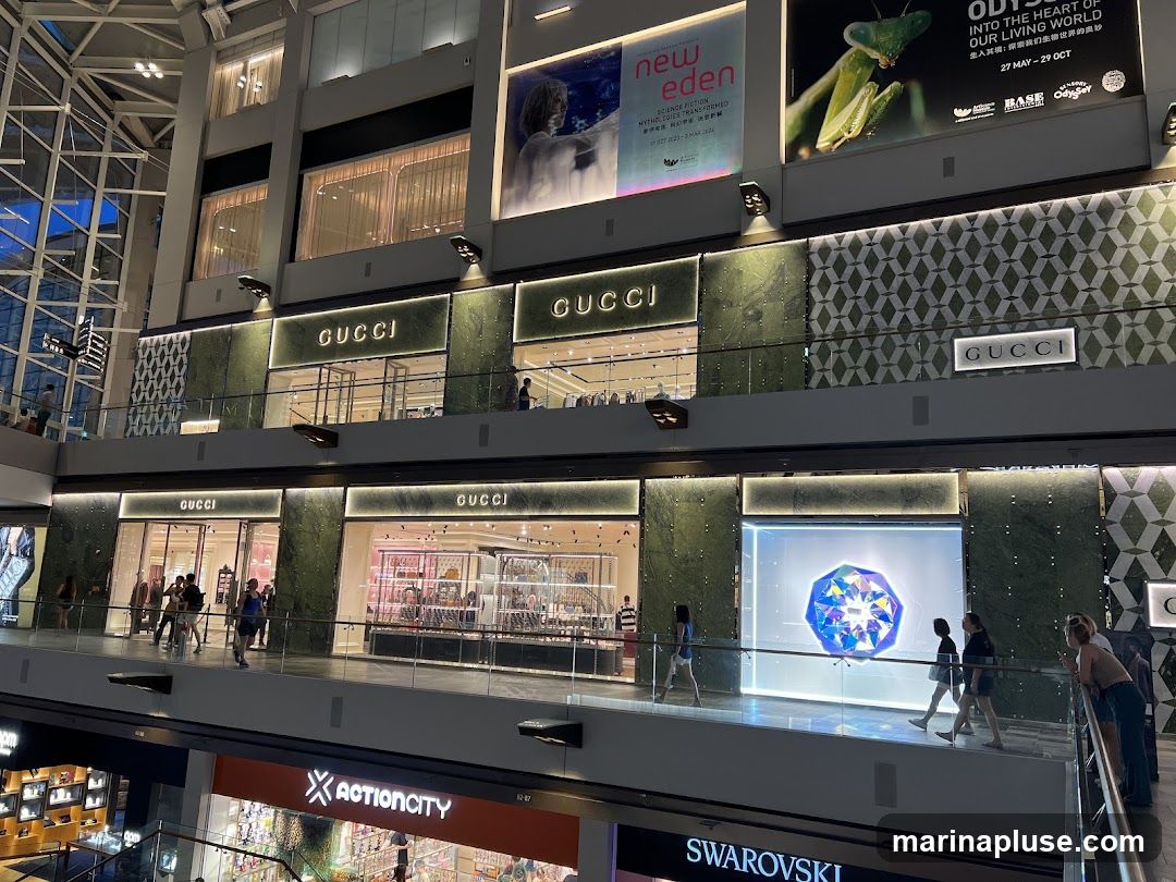 Multiple Gucci storefronts are visible on two levels of a modern shopping mall. The stores feature the Gucci logo prominently above their entrances and display windows. On the upper level, one storefront has a large display window showing a faceted, glowing gem. On the lower level, several storefronts are visible, with people browsing inside and walking past. Other store signs like Actioncity and Swarovski are also visible below. Large advertisements are displayed on the upper walls of the mall.